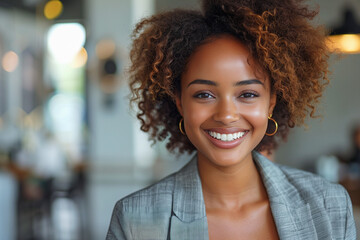 A woman with curly hair smiles brightly at the camera in a coffee shop. She is wearing a gray blazer and gold hoop earrings