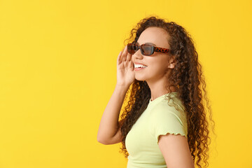 Happy young African-American woman in sunglasses on yellow background