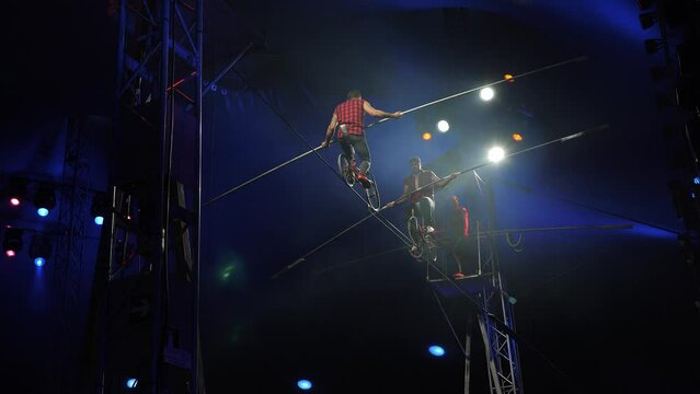Two acrobats ride a bicycle on a tightrope under the dome of the circus. Performance of aerial gymnasts-tightrope walkers in a circus under the light of colorful spotlights. A circus show.