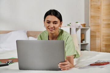 A woman in casual attire lies on a bed, engrossed in her laptop.