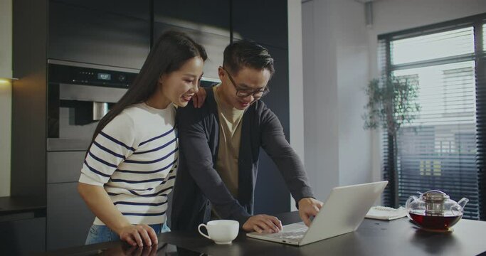 Happy Chinese couple watching video on laptop while standing in modern kitchen. Pretty woman asking questions to her husband and pointing with finger at display. People clinking cups together.