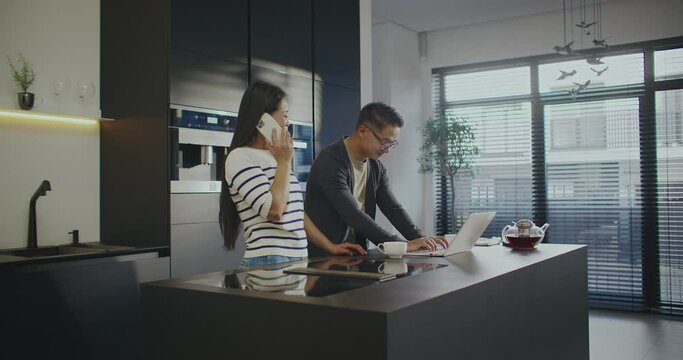 Busy Asian couple using digital gadgets. Young happy woman talking with someone on smartphone while her husband working at laptop. People standing in modern kitchen with panoramic windows.