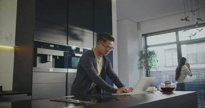 Hardworking freelancer using portative laptop for online work. Woman looking outdoor through window and talking with someone in background. Chinese couple with technology gadgets working from home.