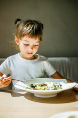 A young girl eating vegetarian pasta with green pesto and black olives at a table, looking cute and content