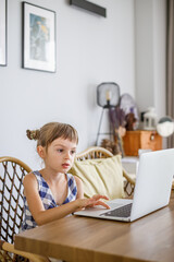A cute 5-6-year-old girl, focused on distance learning, sitting at a wooden table with a laptop