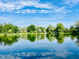 summer lake in the park, blue sky with white clouds and green trees reflection on the lake surface