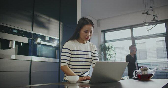 Asian family working remotely from their kitchen. Woman standing at countertop while typing message on her laptop. Male in blurred background walking around and talking on smartphone.