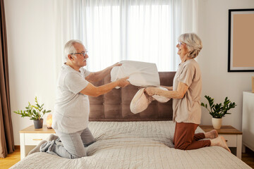 Fun senior couple having pillow fight on a bed in bedroom at cozy home.