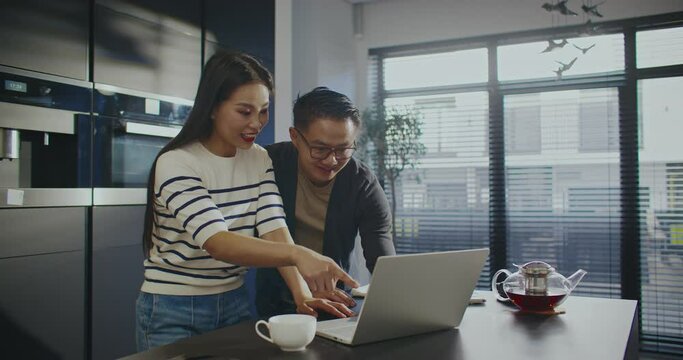 Lovely Asian couple standing in front of laptop. Woman opening lid of portable device. Waving their hands to relatives or friends. Video calling someone. Friendly gesture. Remote communication.