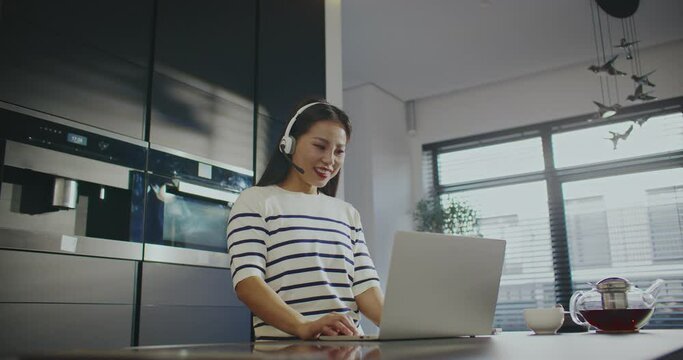 Cute Asian girl standing in front of laptop. Wearing headset with microphone. Talking with potential client or buyer through videocall. Conversation online. Working remotely from home.