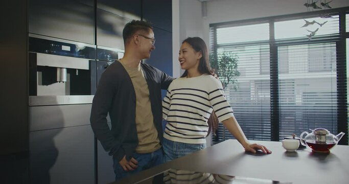 Lovely Asian couple standing in middle of their modern kitchen. Hugging gently. Looking and smiling to each other. Showing love symbol with two fingers to camera. Sun shining bright outside.