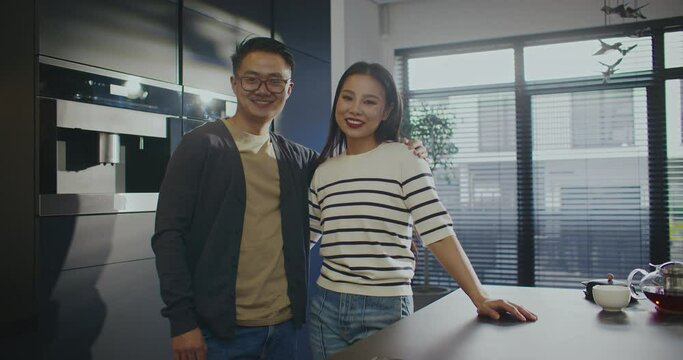 Cute Asian couple standing in middle of kitchen. Hugging each other. Looking directly at camera while smiling. Man with glasses for better vision. Relaxing together during weekend.