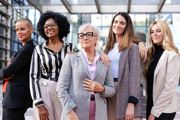 Obraz premium Five businesswomen standing side by side with their arms crossed looking at the camera happy. Suitable for team, friendship and diversity concepts.