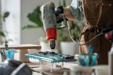 Female artist using dryer to disperse epoxy resin over painting, create drawing of sea waves on canvas. Creative resourceful woman skillfully wields tool to achieve desired effect in art picture.