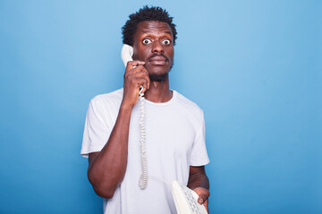Surprised black man with afro hair on blue background talking on a landline phone. Portrait of shocked african american male individual communicating through the telephone.