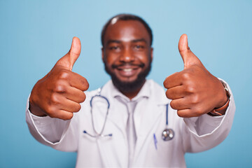 Close-up of cheerful African American doctor posing with his thumbs up towards the camera. Male healthcare specialist standing and gesturing with his hands in front of isolated background.