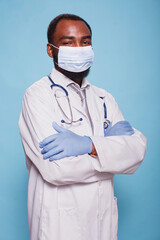 Portrait of healthcare specialist with arms crossed wearing face mask and latex gloves before scrubbing in. Professional medic with stethoscope wearing protective medical gear and white lab coat.