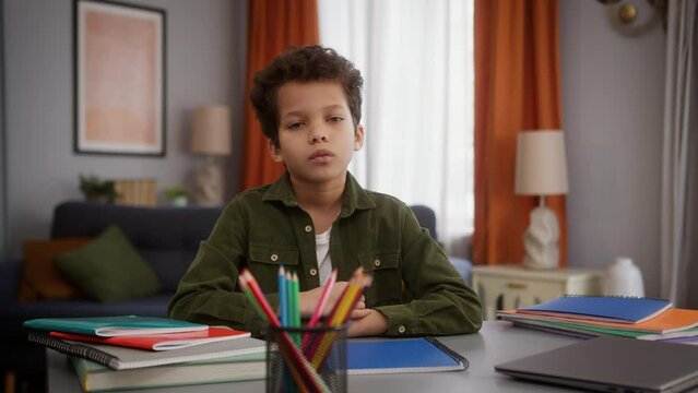 Serious tired sad calm African American ethnic little boy child kid son at home apartment distant education studying at table looking at camera look posing learning study homework childcare health
