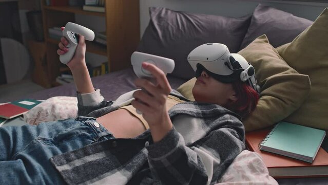 Side shot of red haired gen alpha child immersed in virtual reality, lying in bed with ar headset and using controllers