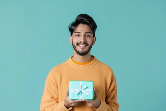 Portrait of a blissful indian man in his 20s holding a gift over soft blue background
