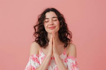 Portrait of a content caucasian woman in her 20s joining palms in a gesture of gratitude over soft pink background