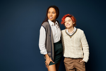 a woman, along with a nonbinary individual, stand together in stylish attire against a dark blue background.