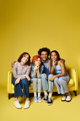 Fototapeta premium A diverse group of young friends, including a nonbinary person, sit and chat comfortably on a vibrant yellow couch in a stylish studio setting.
