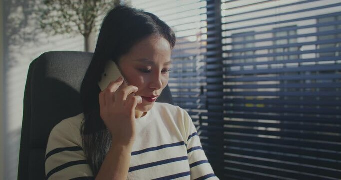 Pretty Chinese girl actively talking on smartphone. Comfortably sitting in chair next to large windows. Having distal conversation. Remotely communicating on phone in office or cosy room.