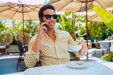 Smiling young stylish businessman using smartphone outdoors. Casual man working on terrace of cafe, broadly smiles during a phone call, drinks a coffee.
