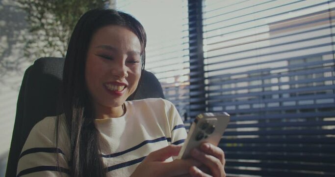 Happy Chinese woman widely smiling while receiving new message on phone. Having positive emotions when typing answer. Chatting in social media. Sitting in front of large window with drapes.