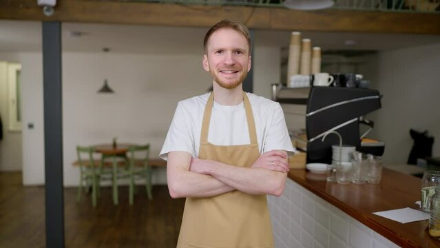 Portrait of a happy blond guy waiter with a beard who crosses his arms on his chest and poses in a cafe