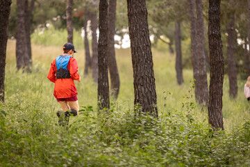 Fototapeta premium A woman is running through a forest with a blue backpack on. The forest is lush and green, and the woman is wearing a red jacket