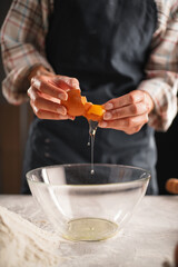 Close-up of female hands separating an egg yolk from the white over a clear bowl in the kitchen. The person is wearing a plaid shirt and a dark apron,