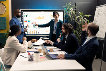 Public relations group creating marketing ad campaigns and promotional activities in a conference room, collaborating on development plan. Specialists reviewing sales trends on interactive board.