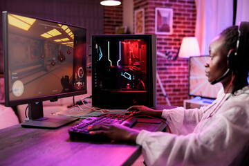 Woman in brick wall living room playing SF video game on gaming PC at computer desk, winding down after work. Gamer girl challenging other players in online multiplayer futuristic shooter