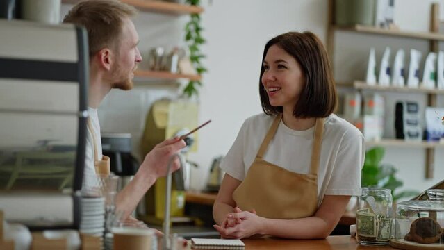 Happy brunette girl in a white T-shirt a cafe worker stands behind the counter talking to a guy barista while drawing up his plan in a cafe
