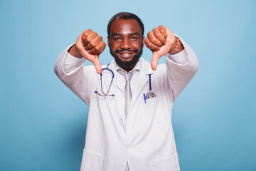 Naklejka premium Black man in hospital uniform and stethoscope stands and gives the camera thumbs down. Hand gestures of disapproval are made by African American healthcare practitioner in this portrait.