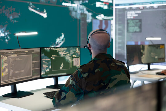 IT engineer operator examines surveillance footage from satellite system, using his computer and a big screen in military command center. Soldier collecting intelligence for missions.