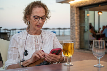 Senior Caucasian woman drinking beer and using smartphone at beachside bar in the Mediterranean