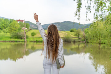 A woman with long hair is standing by a lake, holding her hand up in the air. The scene is peaceful and serene, with the woman's pose and the calm water creating a sense of tranquility.