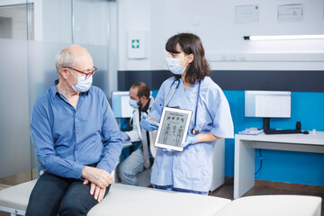 Obraz premium Female practitioner describes human skeleton on a tablet to a senior patient wearing a face mask. Retired old man receiving a medical diagnosis from nurse holding digital device.