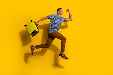 Full length photo of cheerful positive guy dressed print shirt jumping holding luggage running empty space isolated yellow color background