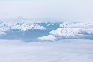 Aerial view of fluffy white clouds and snowy peaks. Snow covered mountains as seen through window of an aircraft flying above clouds