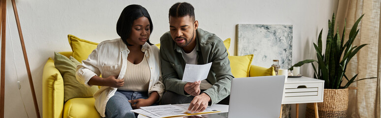 An African American couple sits on a yellow sofa, reviewing paperwork and using a laptop. © LIGHTFIELD STUDIOS
