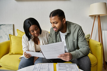 A loving African American couple sits on a yellow couch, reviewing paperwork together.