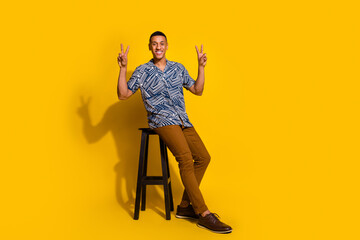 Fototapeta premium Full length photo of cheerful positive guy dressed print shirt sitting stool showing v-signs isolated yellow color background