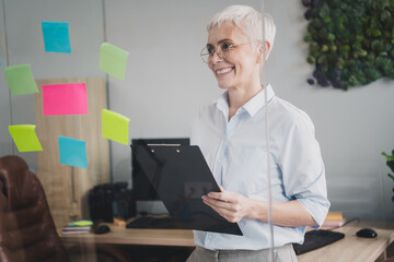 Photo of smiling positive mature lady specialist formalwear writing stickers new ideas indoors workplace workstation