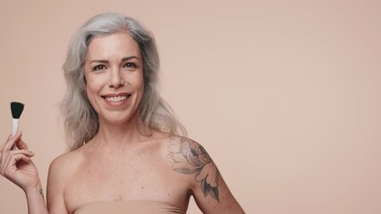 A joyful senior woman elegantly applying makeup with a brush against a soft beige background, exuding ageless beauty and confidence.