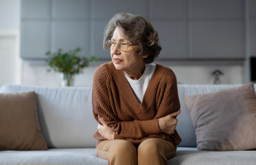 Obraz premium Unhealthy elderly frowning woman sitting on couch, putting hands on belly, suffering from strong stomach ache and having painful feeling in abdomen