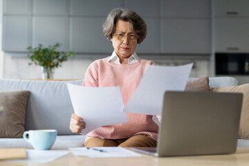 Serious European elderly woman in glasses using laptop computer and documents. Bookkeeping, paying bills and taxes